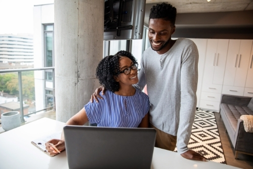 Husband and wife smiling at one another while using laptop