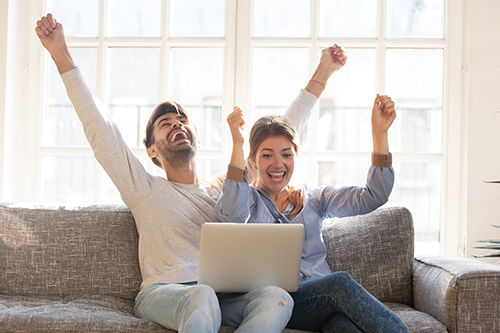 Husband and wife sitting on couch using computer excited with hands in the air